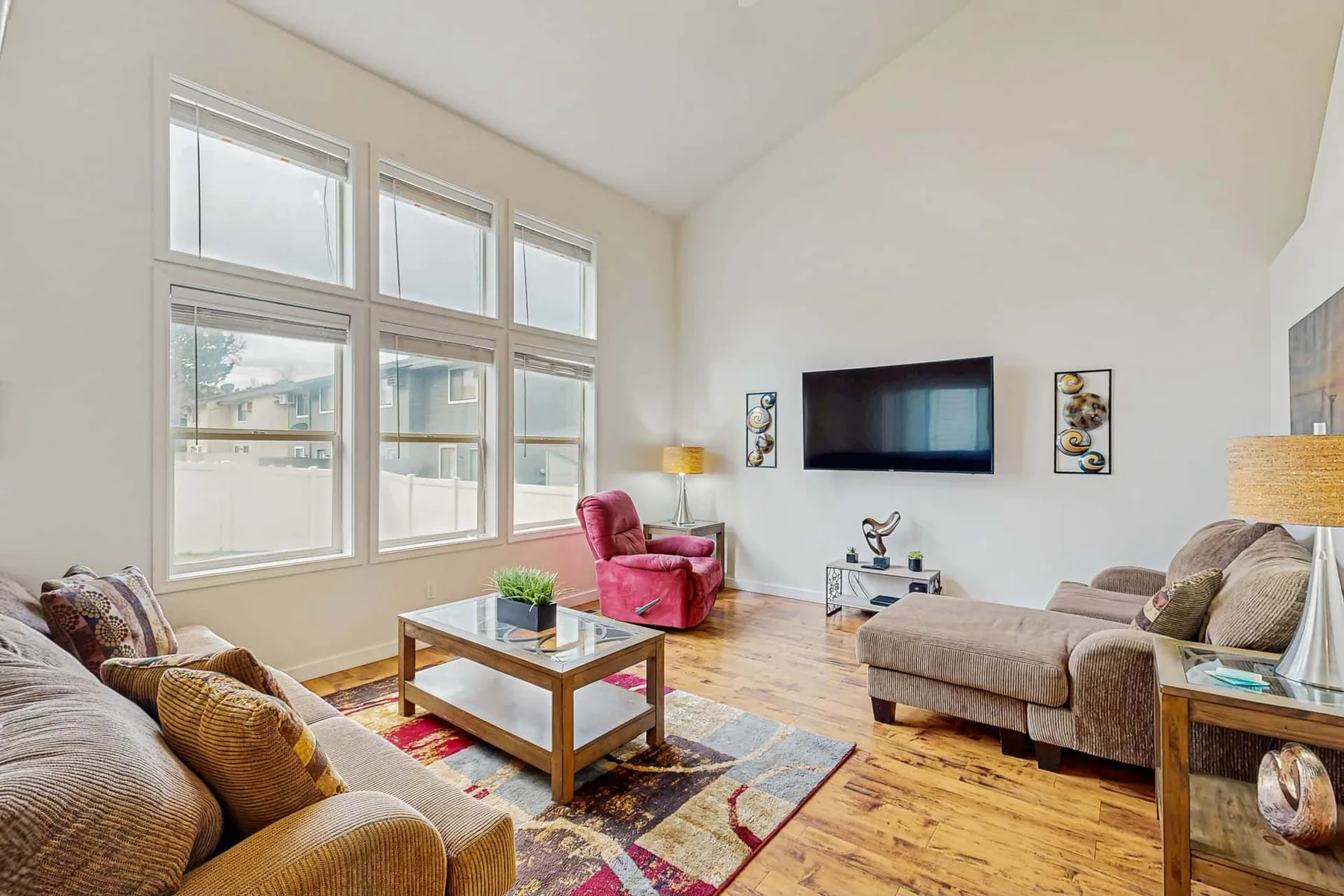 A bright living room with large windows, a wall-mounted television, and seating around a coffee table.
