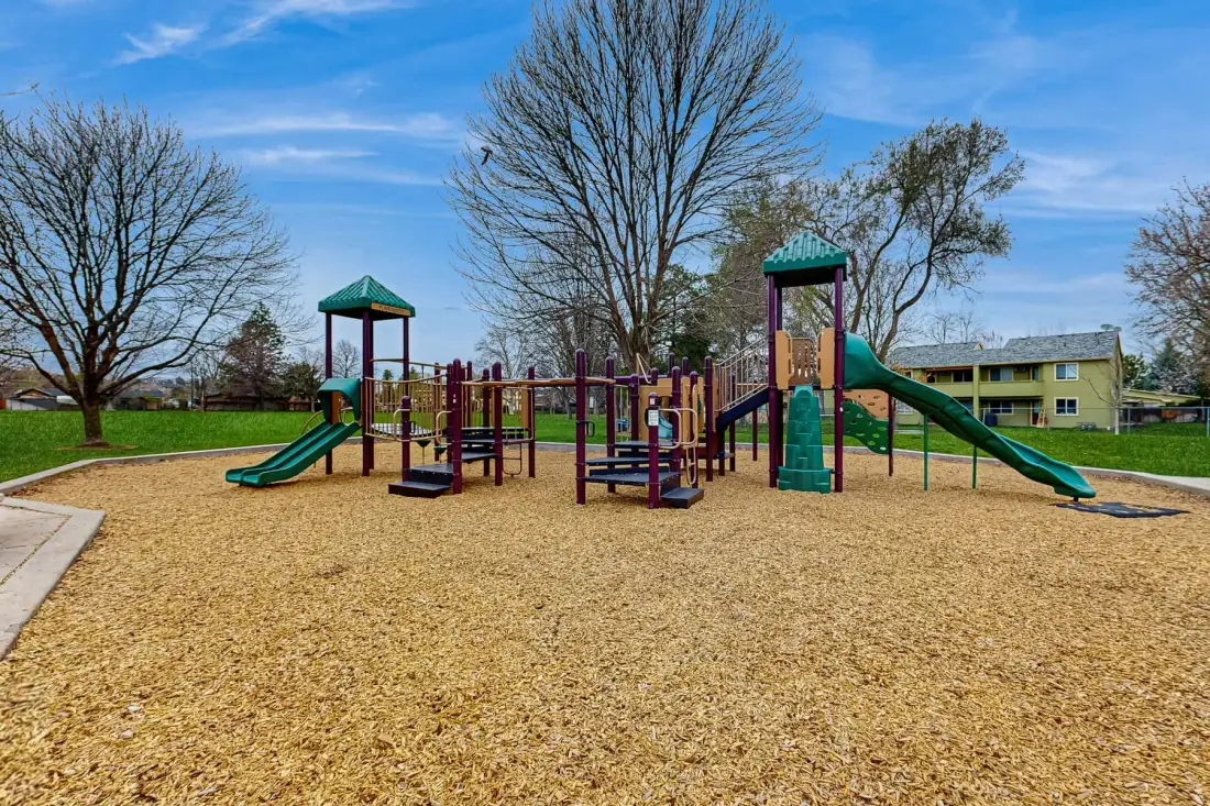 A playground with colorful climbing structures and slides stands on a wood-chip surface.