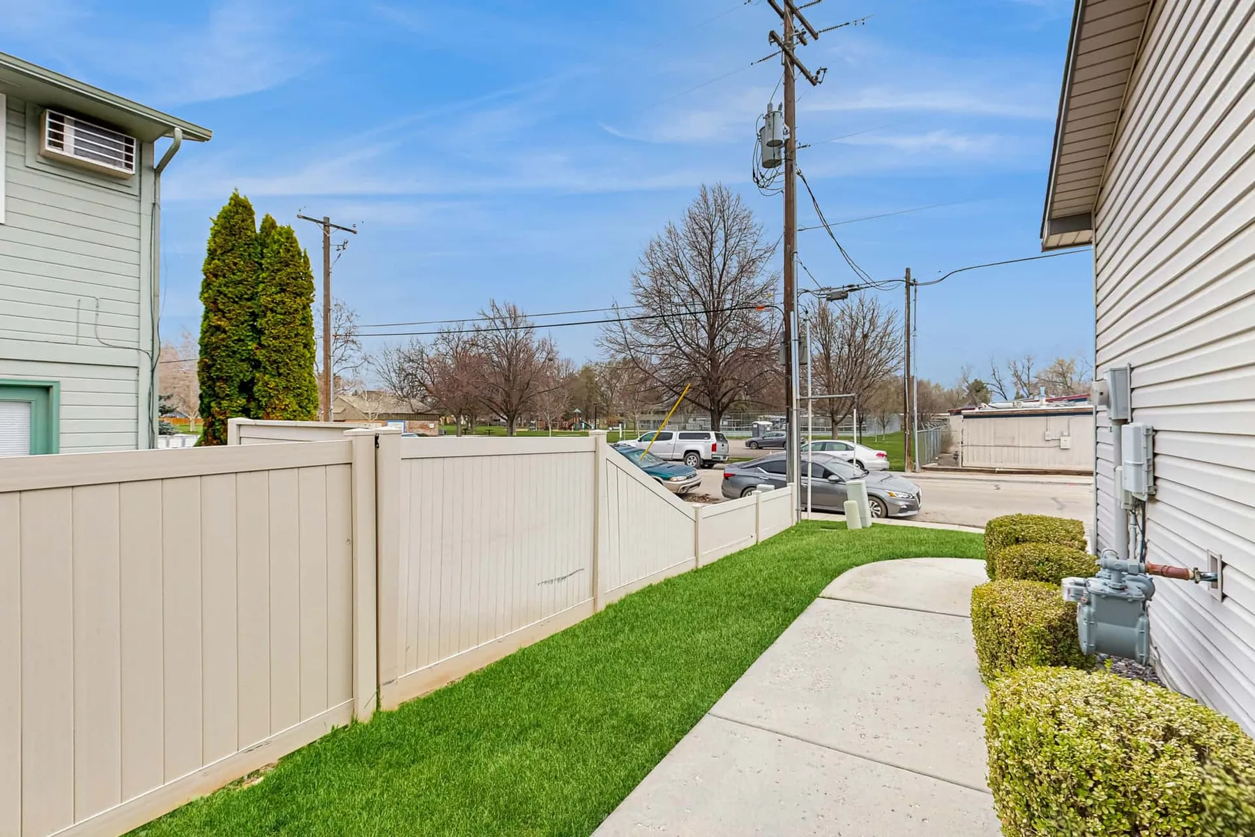 A concrete walkway runs between a light-colored fence and a building, with grass and shrubs alongside.