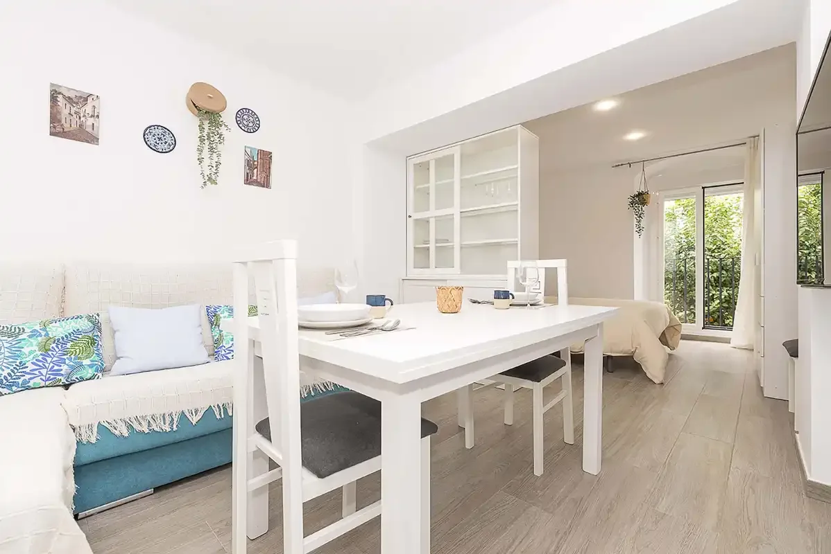 A bright open-plan room with a dining table, seating, and a bed beside glass doors.