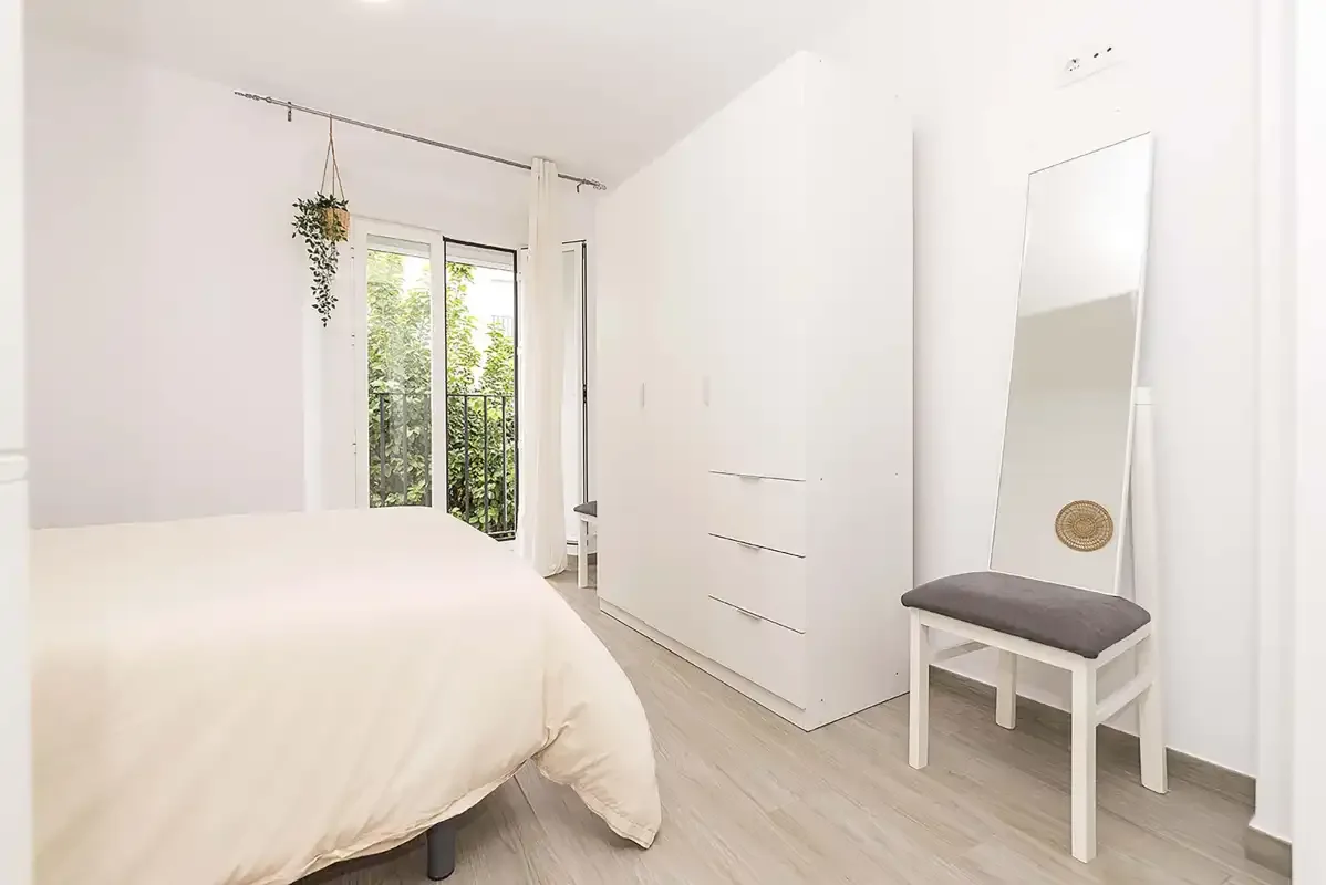 A bright bedroom with a bed, wardrobe, and a chair beside a full-length mirror.