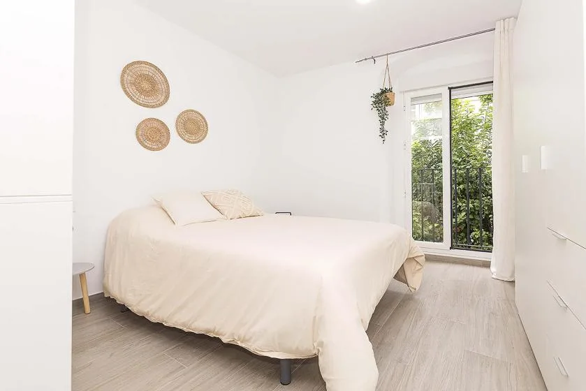 A bright bedroom with a bed beside a tall window and light curtains.