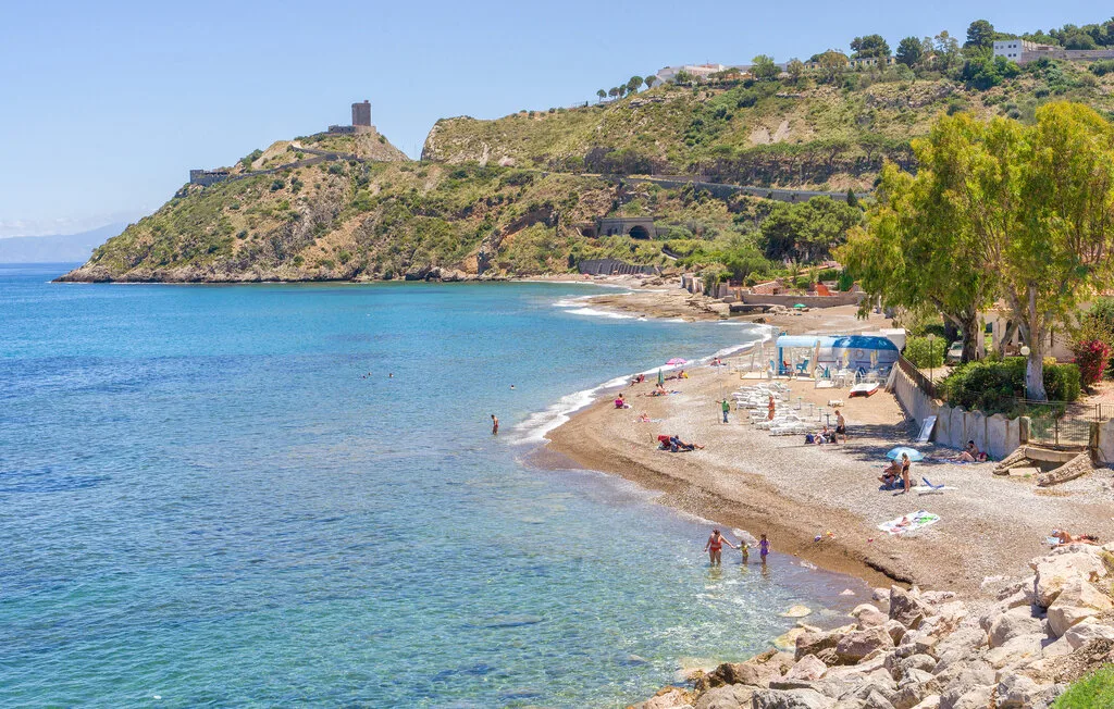 A curved beach borders clear blue water below a green hillside.