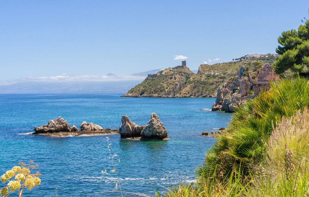 A coastal landscape with blue water, rocky outcrops, and green vegetation.
