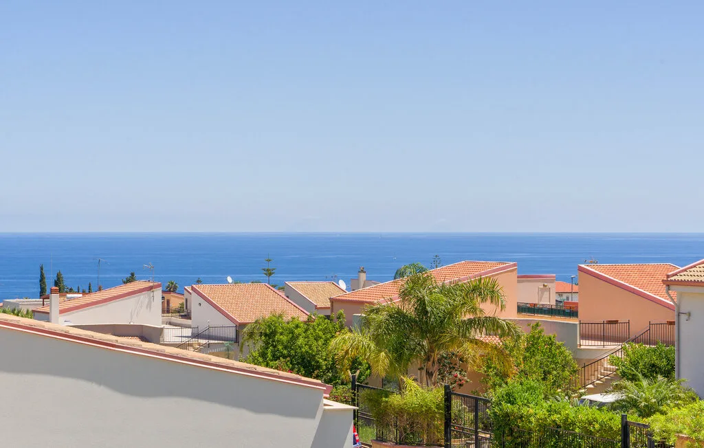 A coastal residential scene with red-tiled roofs, greenery, and the sea beyond.