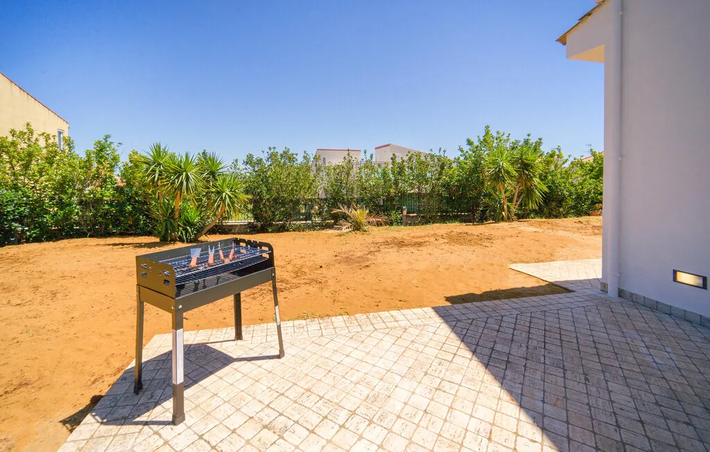 An outdoor patio with a metal grill beside a sandy yard and leafy shrubs.
