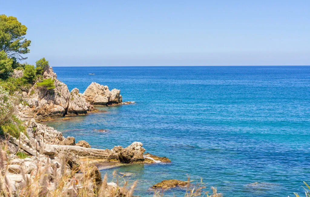 A rocky coastline borders clear blue water beneath a bright sky.