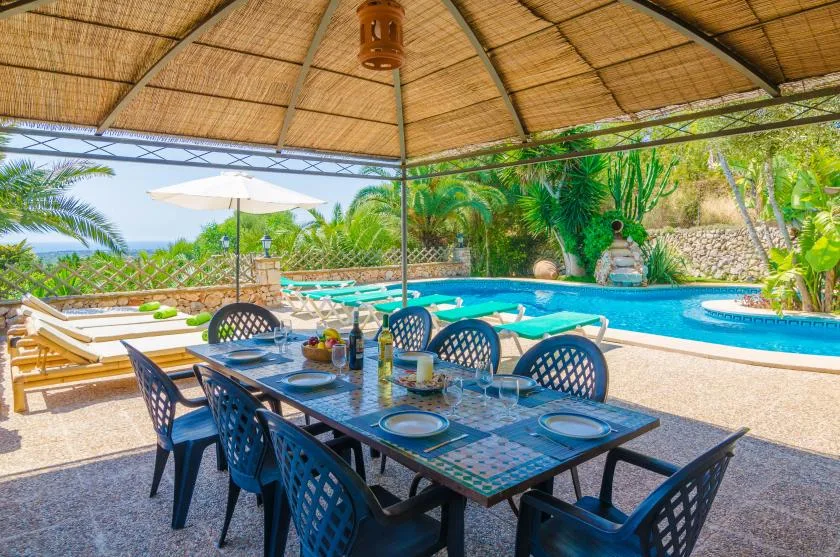 Zona de comedor exterior a la sombra junto a una piscina, con tumbonas cerca.
