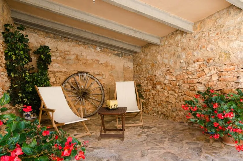 Un patio de piedra con asientos al aire libre, una rueda de madera y plantas con flores.