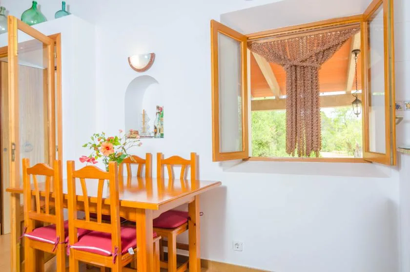 Una zona de comedor con asientos de madera junto a una ventana abierta con vistas a la vegetación.