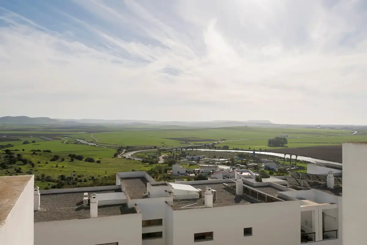 Una vista desde una azotea muestra edificios blancos y campos verdes bajo un cielo nublado.