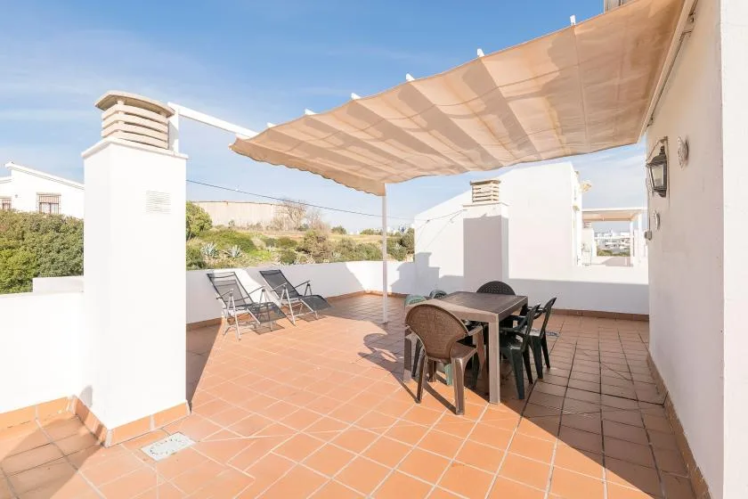 Una terraza soleada tiene una mesa de comedor y sillas bajo un toldo de tela.