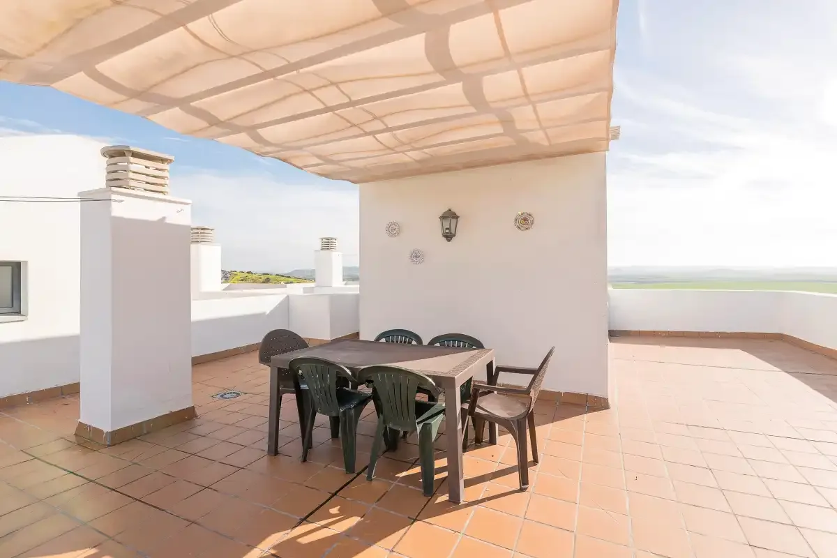 Una terraza sombreada en la azotea tiene una mesa de comedor y asientos junto a una pared blanca.