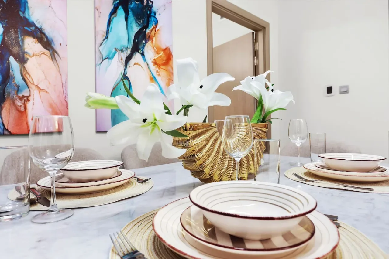 A dining table is set with plates, glassware, and white lilies beneath colorful abstract artwork.