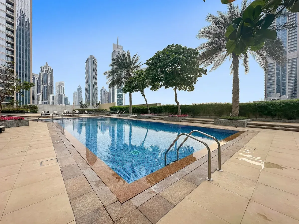 Una piscina al aire libre está rodeada por una terraza pavimentada, palmeras y edificios altos.
