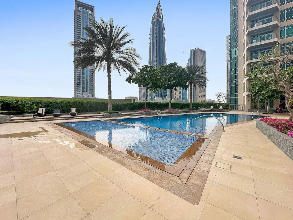 An outdoor swimming pool surrounded by a paved deck, palm trees, and high-rise buildings.