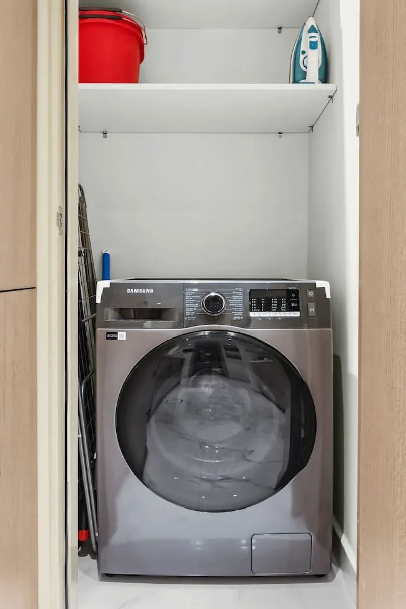 A front-loading washing machine sits in a laundry closet beneath a shelf.