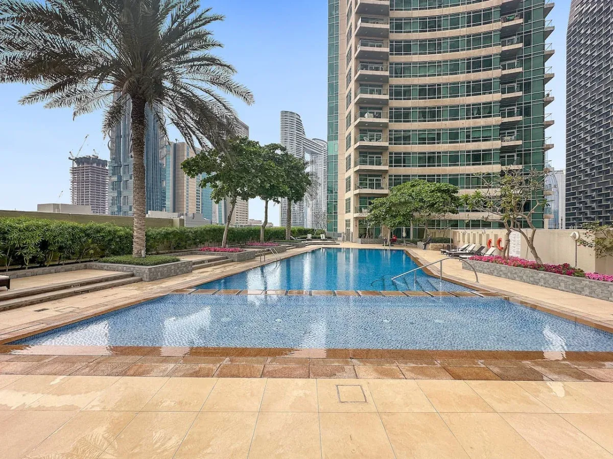 A rooftop swimming pool sits beside a high-rise building, with palm trees and city towers nearby.