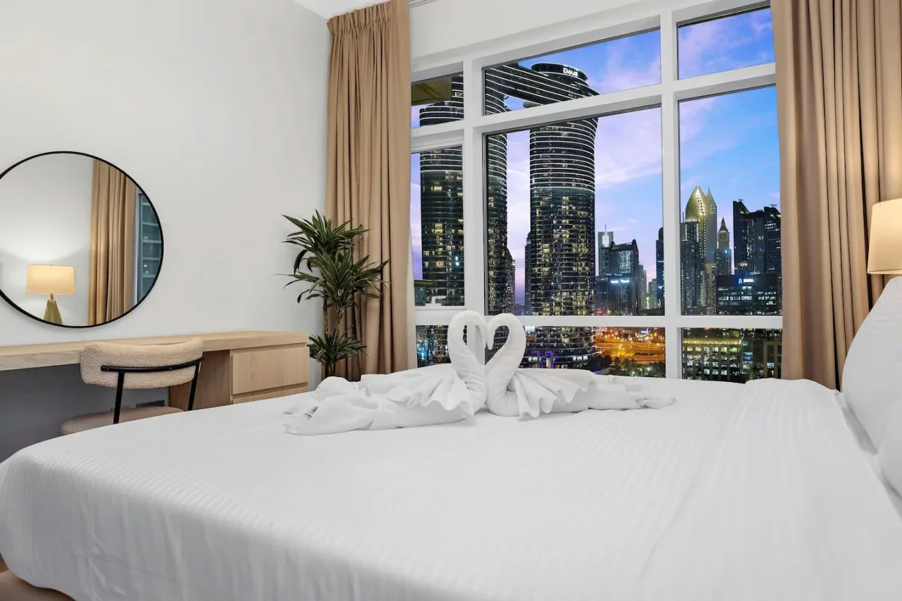 A bedroom with a neatly made bed and a large window overlooking city buildings.