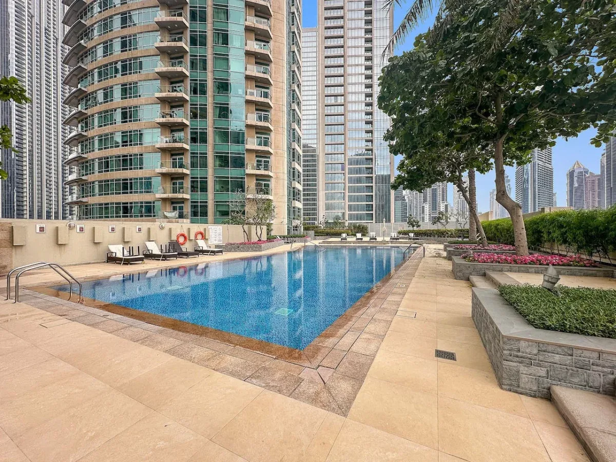 An outdoor swimming pool sits among high-rise buildings, with paved paths and landscaped greenery.