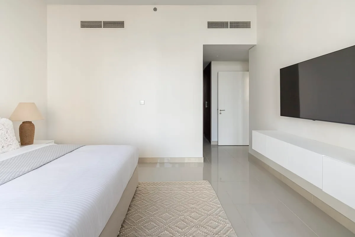 Un dormitorio luminoso con una cama, un televisor montado en la pared y una alfombra clara.