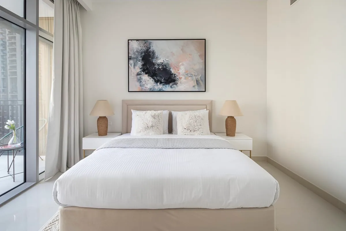 Un dormitorio luminoso con una cama bien tendida y una obra abstracta encima.