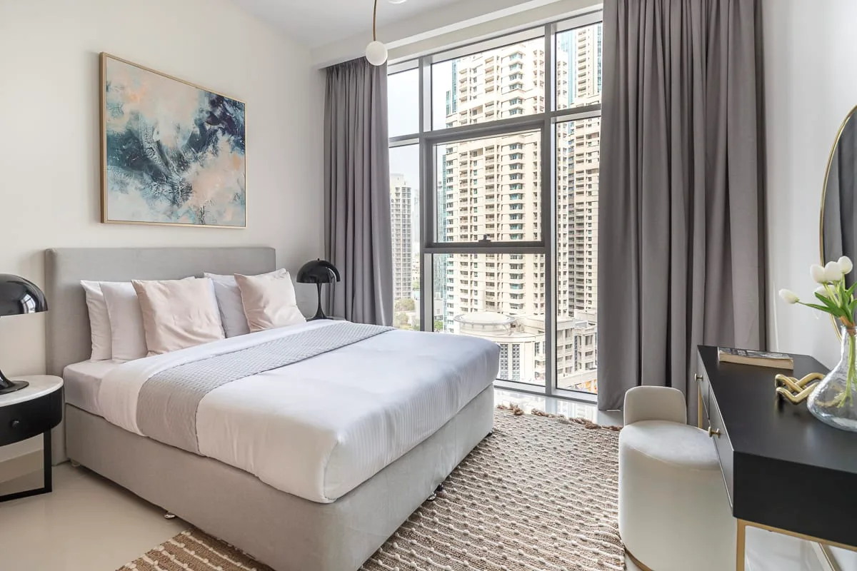 Un dormitorio con una cama tendida junto a ventanales que dan a edificios cercanos.