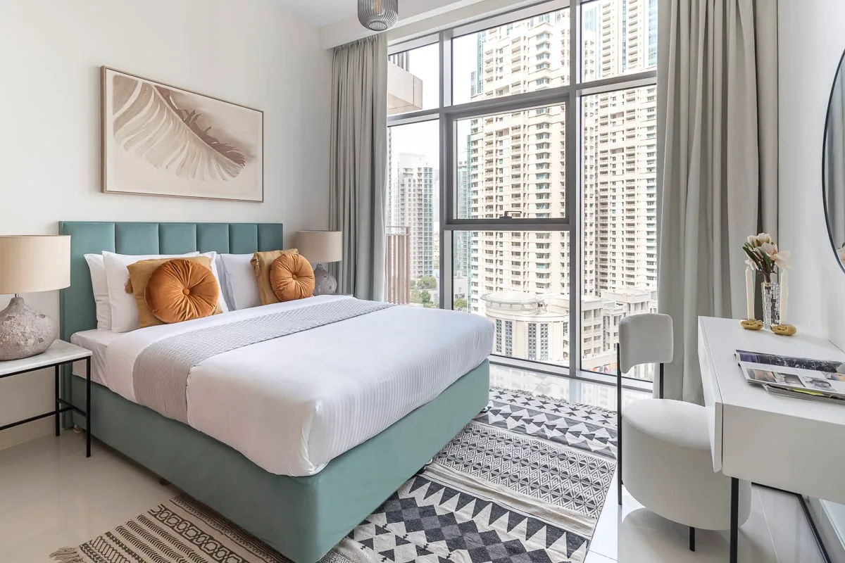 Un dormitorio con una cama bien tendida junto a grandes ventanas con vistas a edificios de la ciudad.