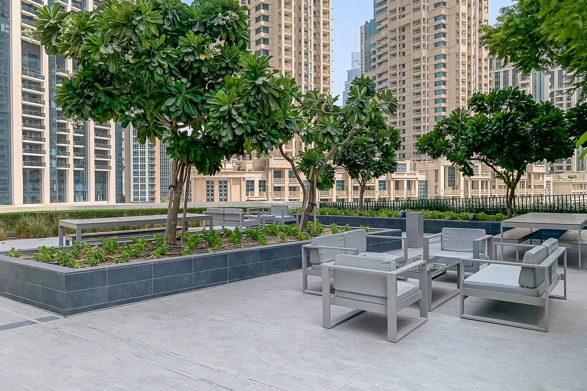 Una zona de estar al aire libre con muebles grises, jardineras y árboles entre edificios altos.