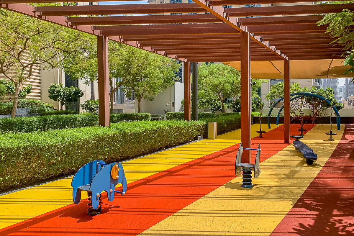 Un colorido parque infantil al aire libre bajo una pérgola de madera, con juegos y setos.
