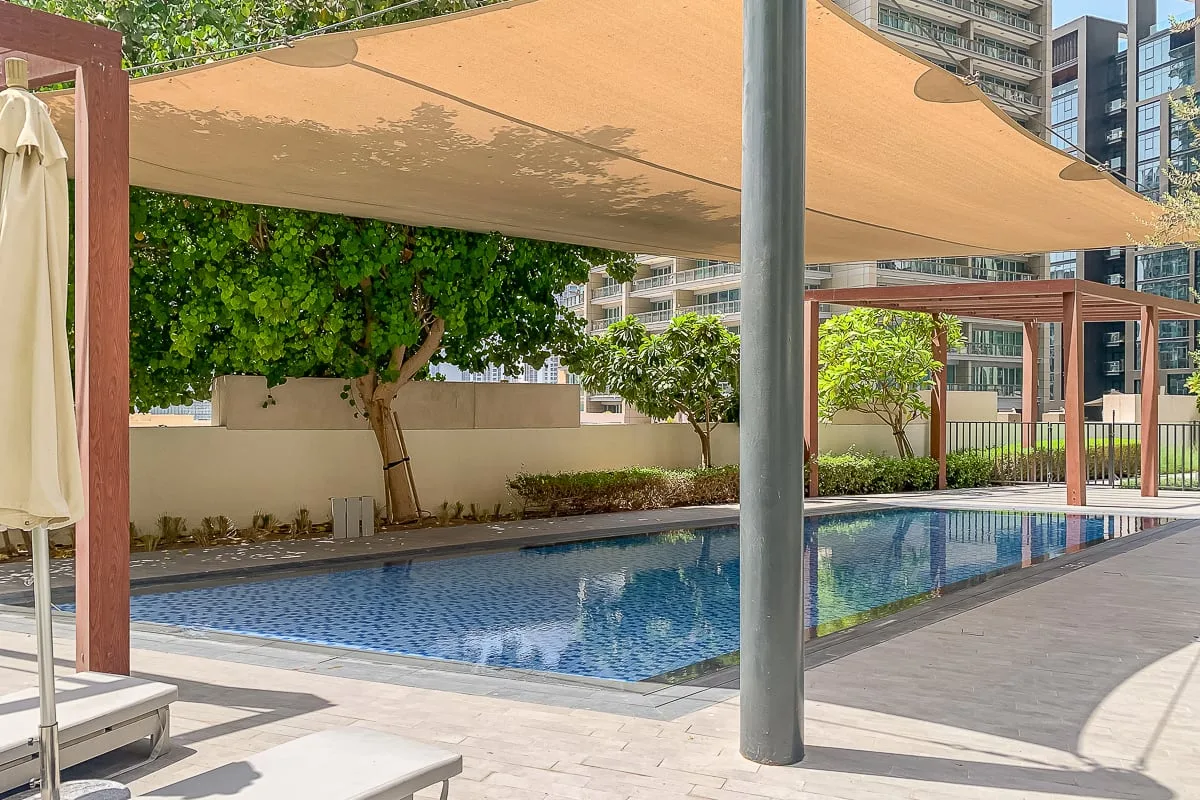 Una piscina al aire libre está bajo una gran cubierta de sombra, con árboles cerca.