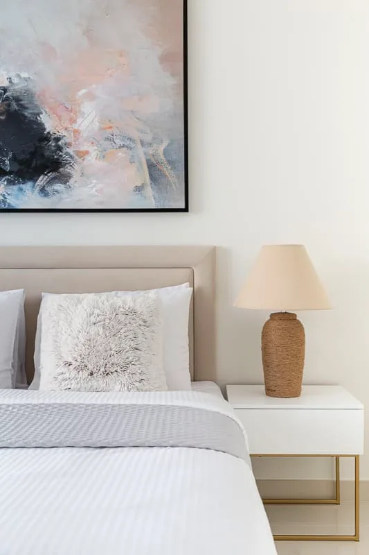 Una cama bien arreglada está junto a una mesita con una lámpara y una obra enmarcada encima.