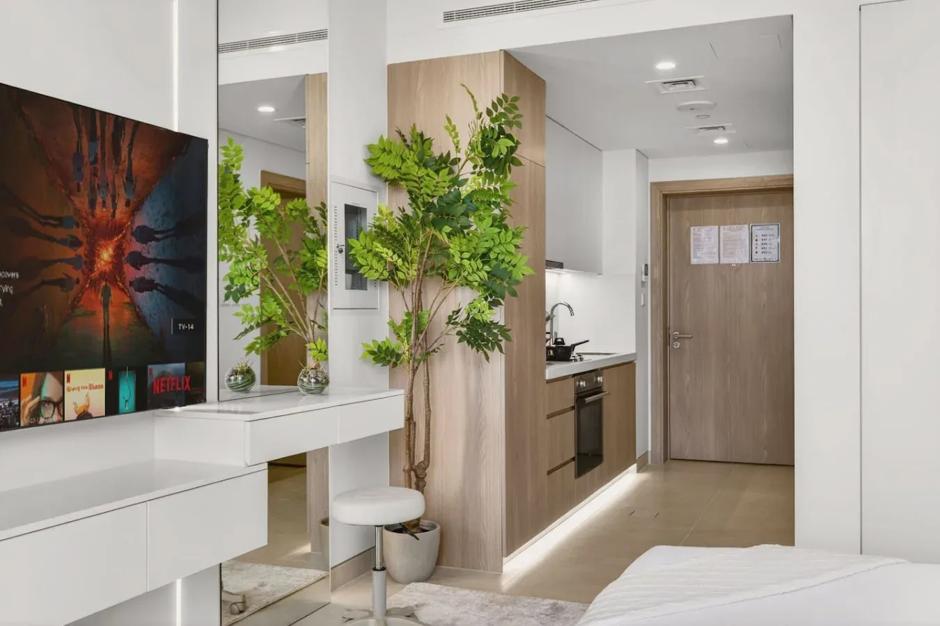 A bright modern bedroom opens onto a compact kitchenette, with a vanity and wall-mounted television.