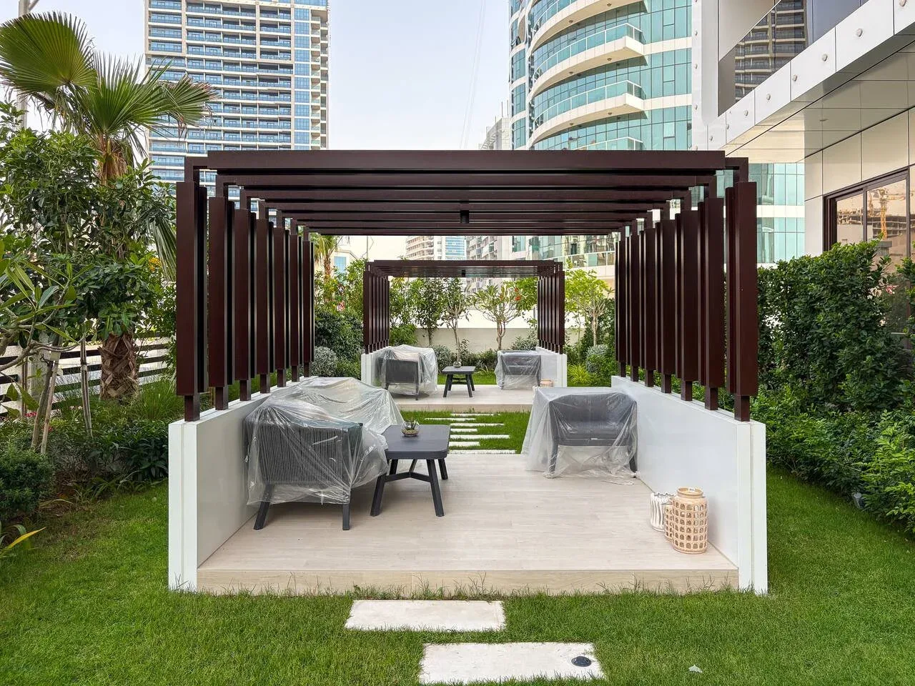 An outdoor seating area beneath a dark pergola, surrounded by greenery.