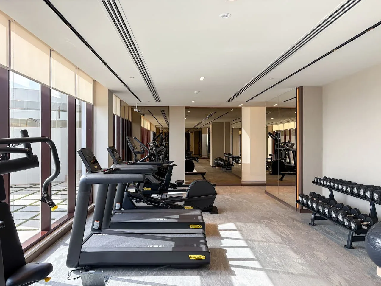 A fitness room with treadmills, exercise machines, and dumbbells beside large windows.