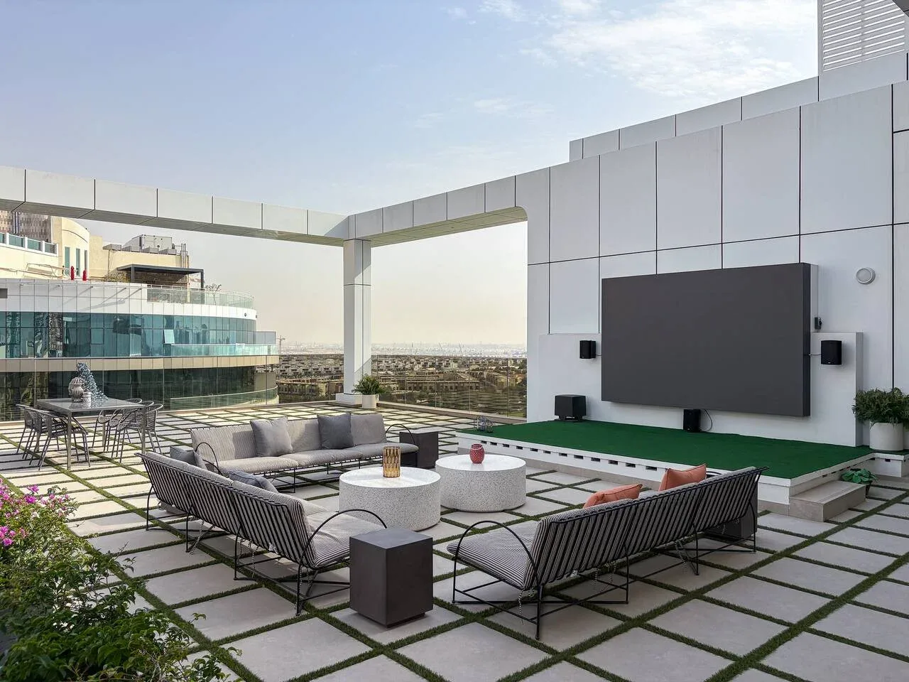 A rooftop terrace lounge has outdoor seating arranged around low tables beside a large screen.