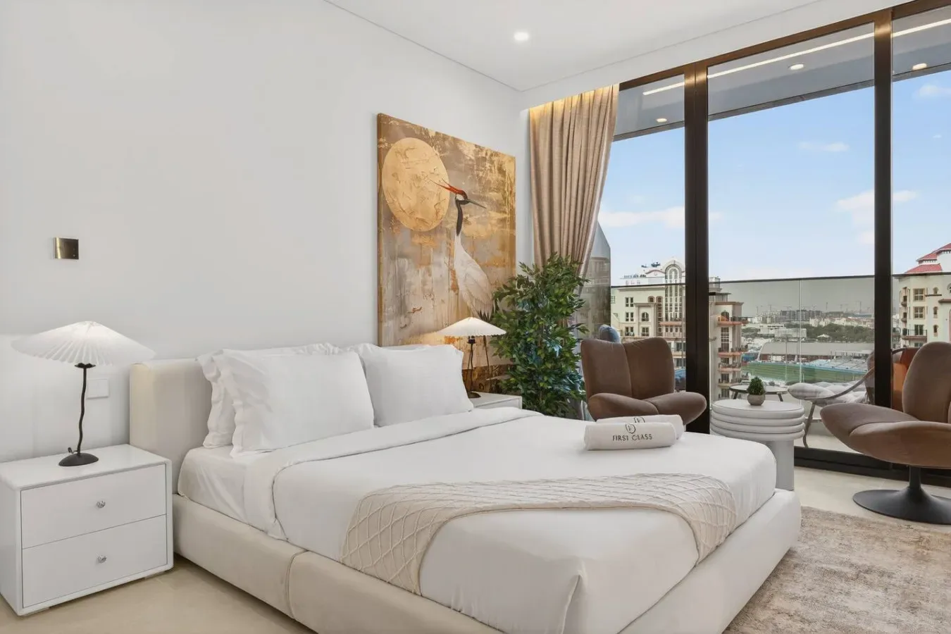 A bright bedroom with a white bed beside floor-to-ceiling windows.