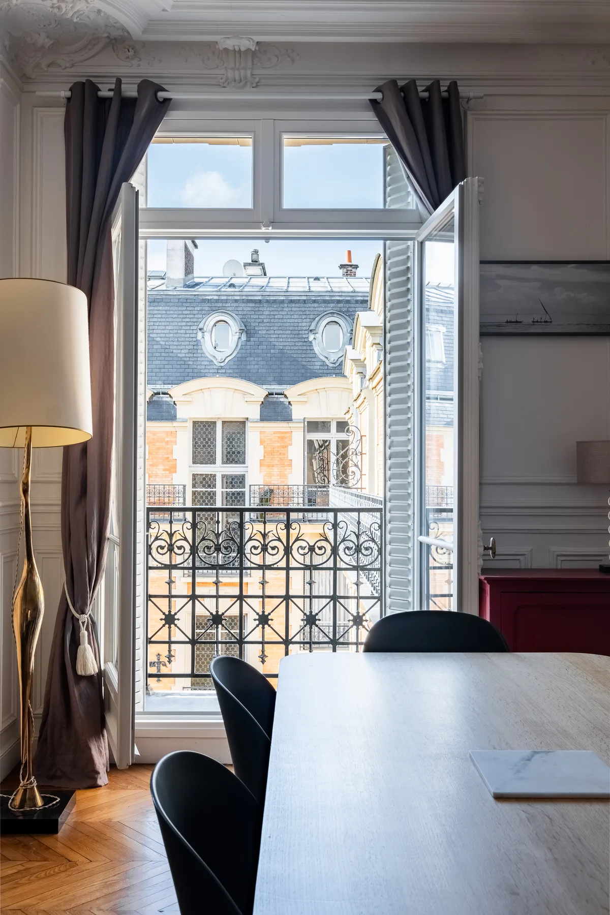 A dining area with a table and chairs beside open windows overlooking nearby buildings.