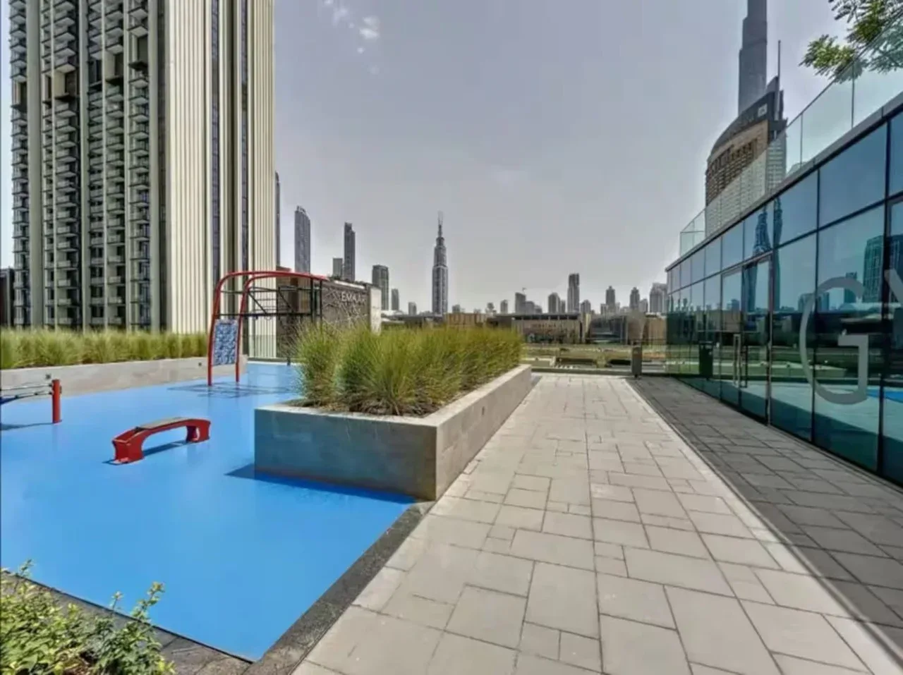 Una terraza exterior pavimentada con jardineras, suelo azul de juegos y edificios de la ciudad al fondo.