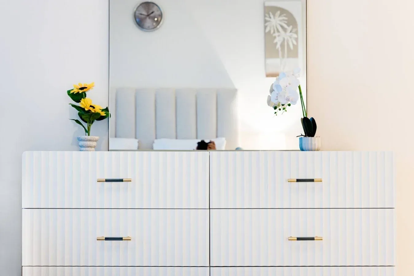 Un dormitorio luminoso con una cómoda blanca, flores y un espejo de pared.