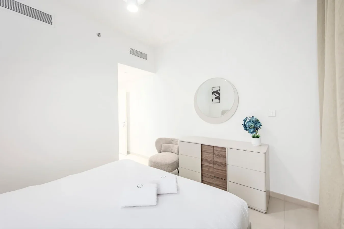 Un dormitorio luminoso cuenta con una cama, una cómoda y un espejo redondo en la pared.