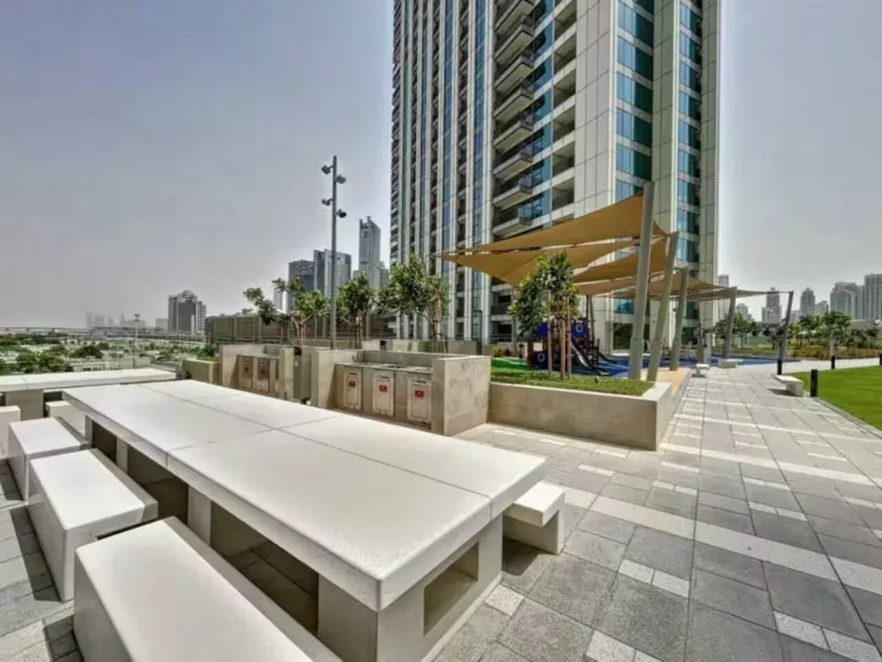 Una terraza exterior pavimentada tiene asientos integrados junto a una zona ajardinada y un edificio alto.