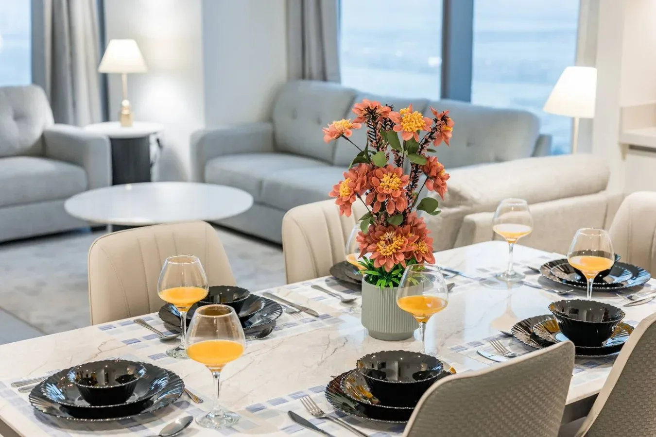 Una mesa de comedor puesta con platos y flores, junto a asientos y grandes ventanas.