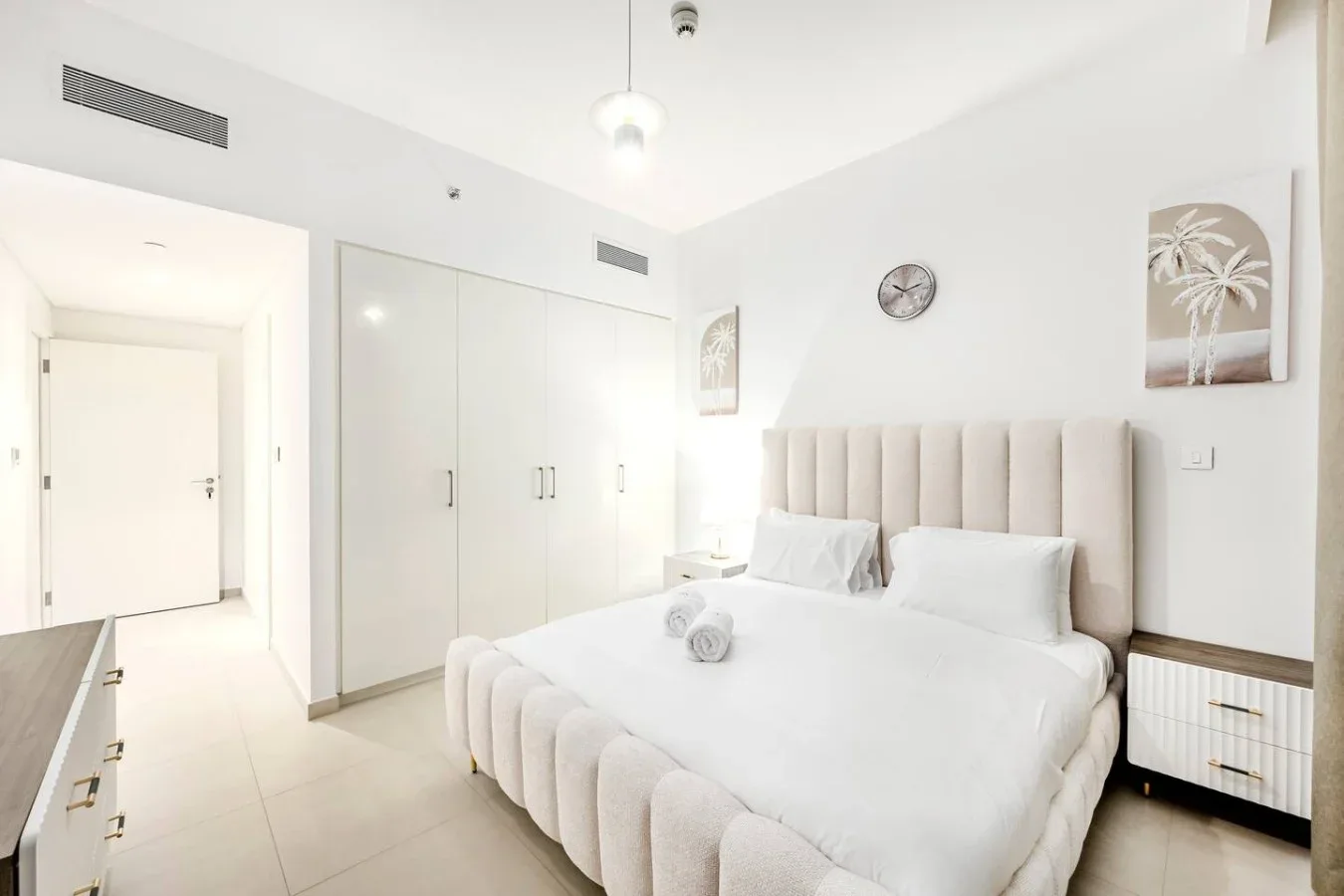 Un dormitorio luminoso con una cama blanca, cabecero tapizado claro y armarios empotrados.