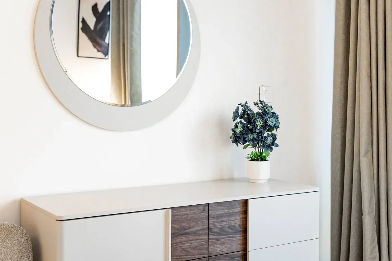 Un rincón luminoso de dormitorio con cómoda, espejo redondo y planta en maceta.