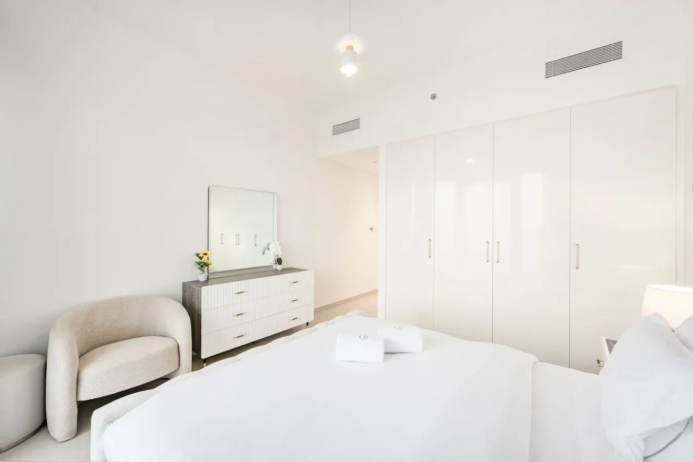 Un dormitorio luminoso con una cama, un asiento tapizado y armarios blancos.