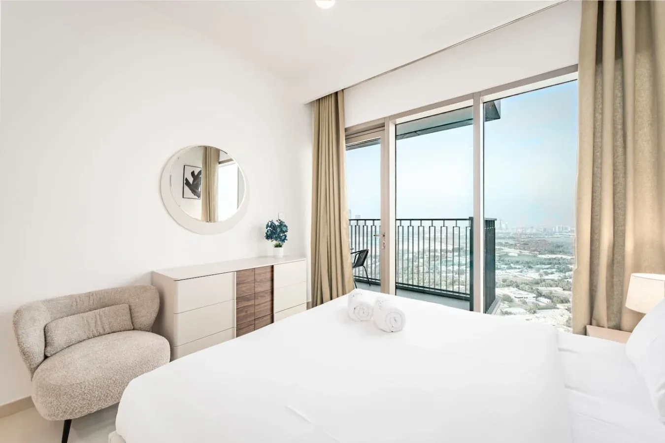 Un dormitorio luminoso con cama, asiento y grandes ventanales con vistas a la ciudad.