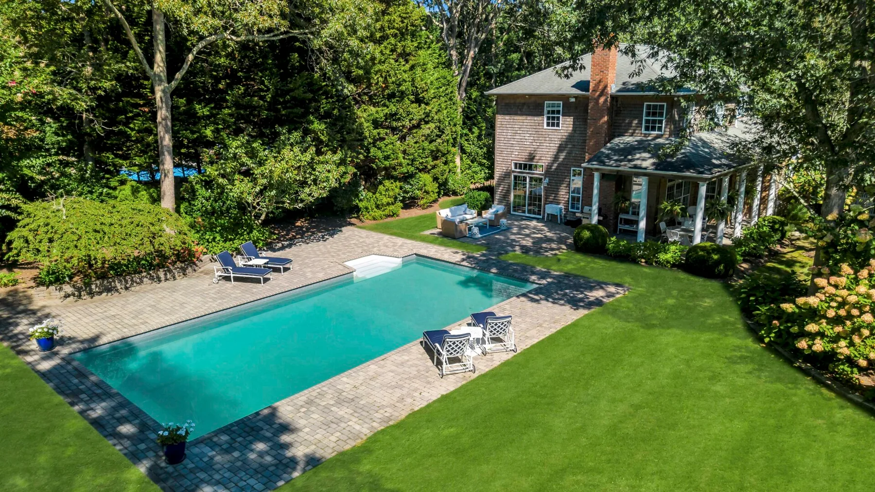 Una piscina y un patio pavimentado están junto a una casa y un césped verde.