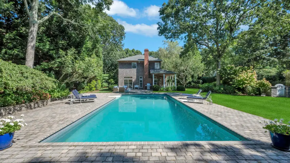 Una piscina al aire libre con una casa de ladrillo y un jardín frondoso al fondo.