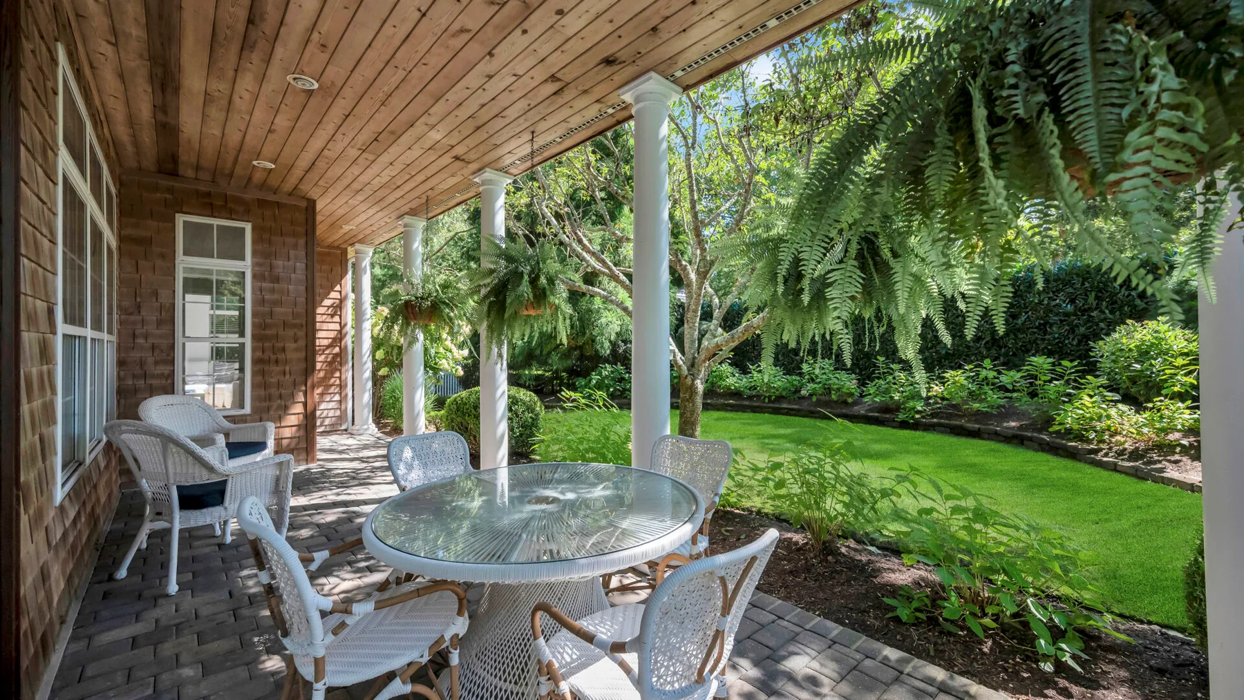 Un patio cubierto con una mesa de vidrio da a un césped verde y un jardín.